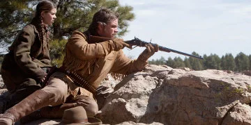 Mattie Ross Sat Behind LaBoeuf Who Is Aiming His Gum Over A Wall Of Rocks